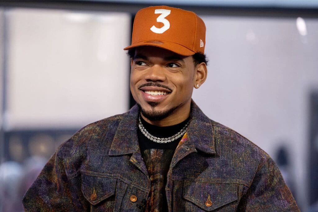 A man wearing an orange baseball cap with the number 3, a patterned jacket, and a diamond necklace smiles while looking slightly to the side.