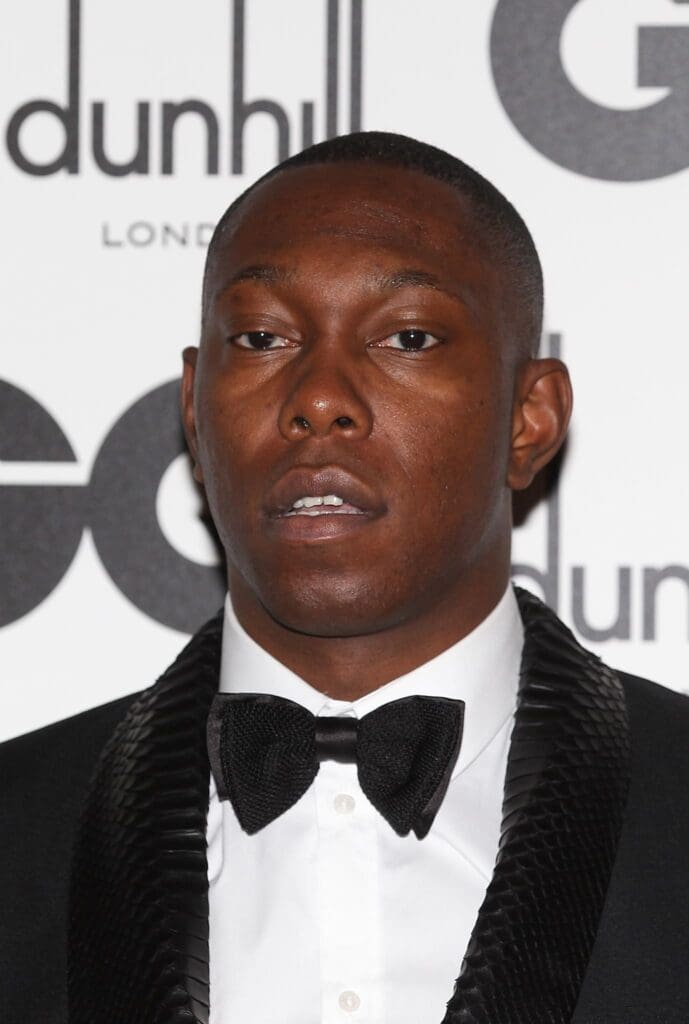 A man in a tuxedo with a black bow tie poses in front of a step-and-repeat backdrop with "dunhill LONDON" and "GQ"—the British rapper and music artist Dizzee Rascal commands attention.