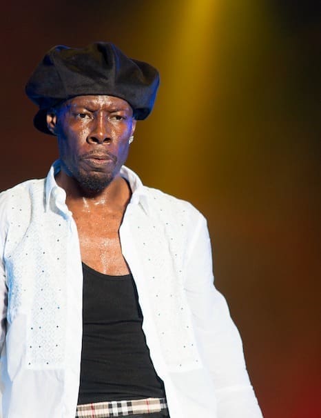 A man wearing a black beret, white shirt, and black tank top performs dancehall on stage under yellow lighting, channeling the vibrant energy of reggae icons like Shabba Ranks.